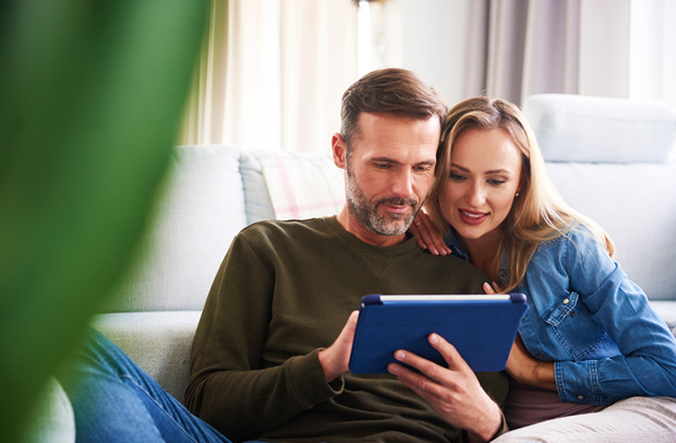 Middle-aged couple on couch reviewing insurance discounts on a tablet.